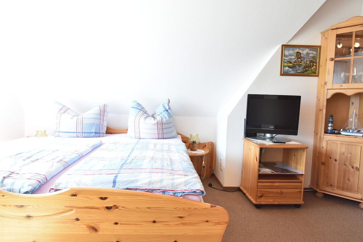 Une chambre à coucher avec un lit, une tête de lit en bois, un téléviseur posé sur un meuble en bois, une petite armoire et un tableau accroché au mur.