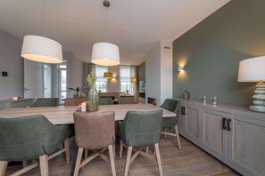 A modern dining area with a wooden table, green upholstered chairs, and pendant lighting. - Luxury 8-person Wellness Villa With Sauna and Outdoor hot tub is Unique (Colijnsplaat)