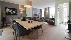 A modern living room with a wooden dining table, black leather chairs, and a grey sofa. There is a TV mounted on the wall and a large window with curtains. - Luxury 8-person Wellness Villa With Sauna and Outdoor hot tub is Unique (Colijnsplaat)