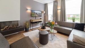 A modern living room with a fireplace, a flat-screen TV, a sofa, a coffee table, and a large window with curtains. - Luxury 8-person Wellness Villa With Sauna and Outdoor hot tub is Unique (Colijnsplaat)