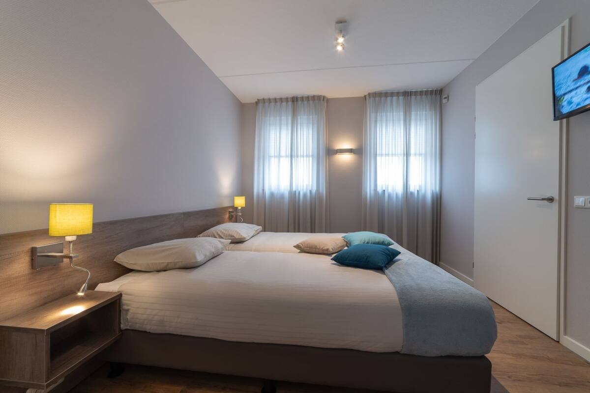 A hotel room with a large bed, two bedside tables with lamps, a flat-screen TV mounted on the wall, and a window with sheer curtains.