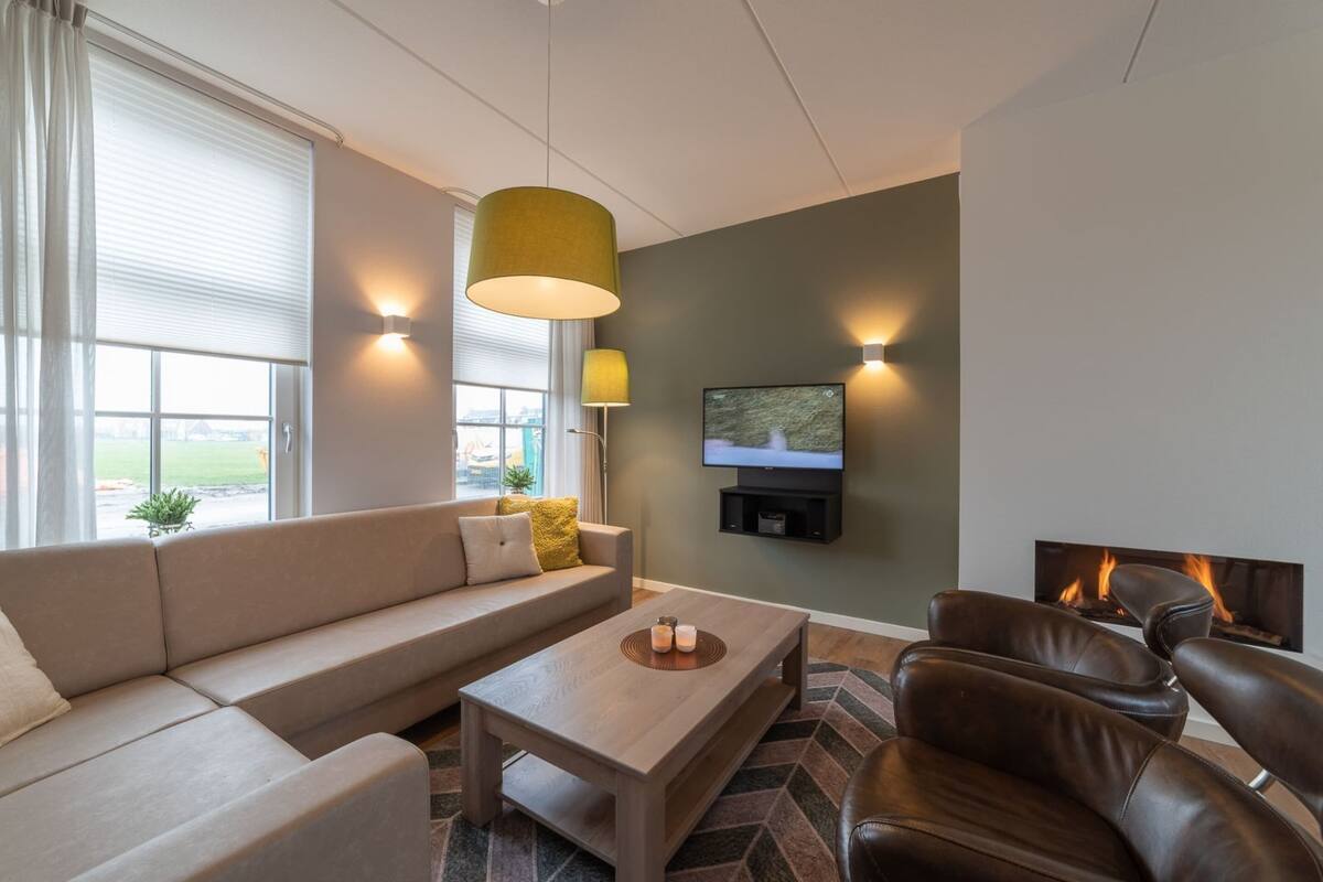 A modern living room with a sofa, coffee table, and leather chairs. A flat-screen TV and a fireplace are visible.
