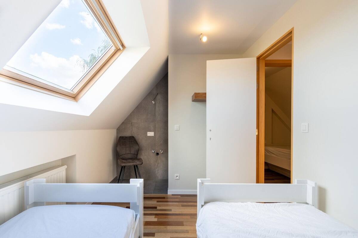 A modern bedroom with two beds, a skylight, a chair, and a staircase leading to a loft.