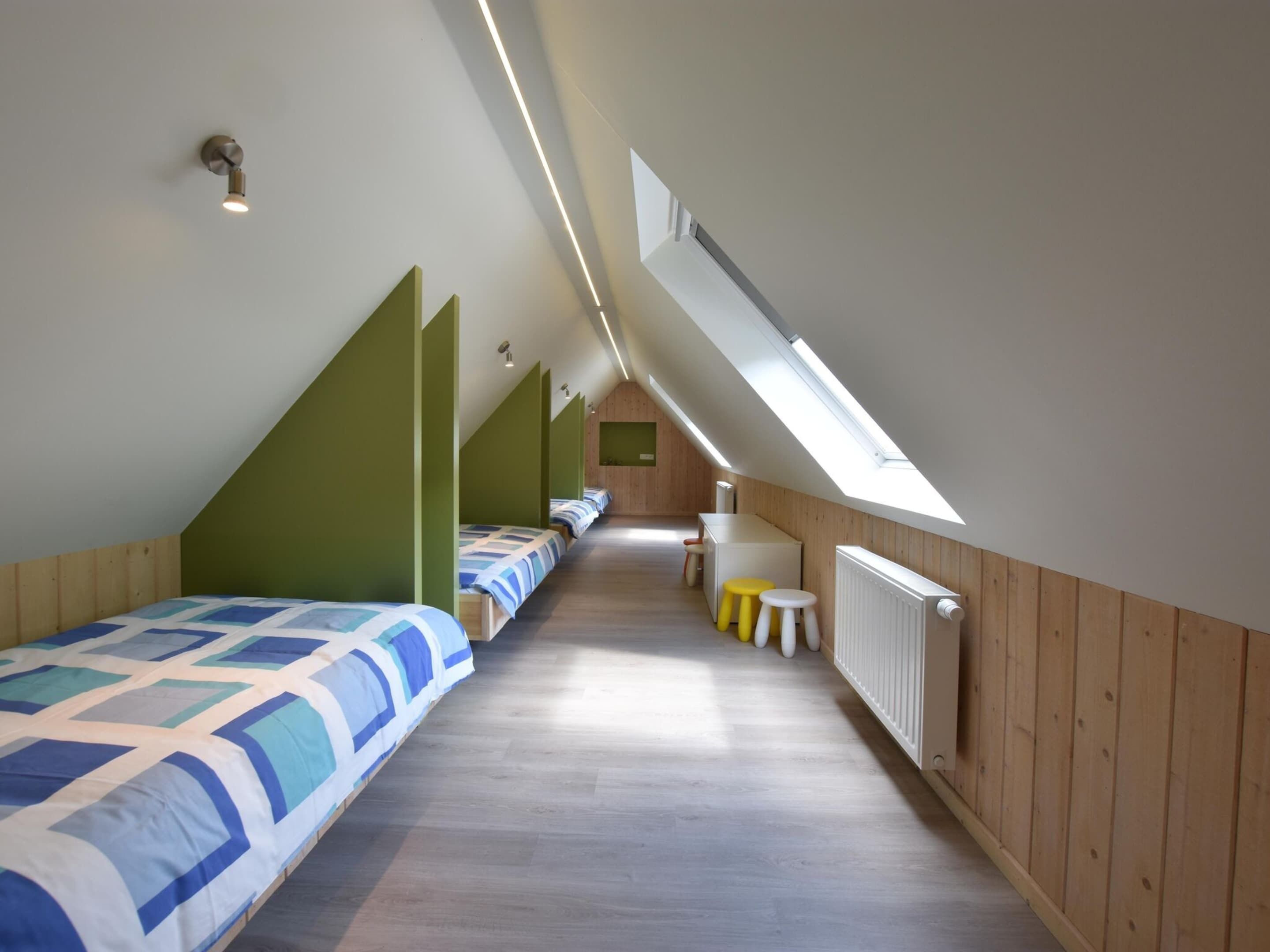A long, narrow room with wooden walls and a sloped ceiling, featuring a row of beds with blue and white bedding, a small table with stools, and a radiator on the right.