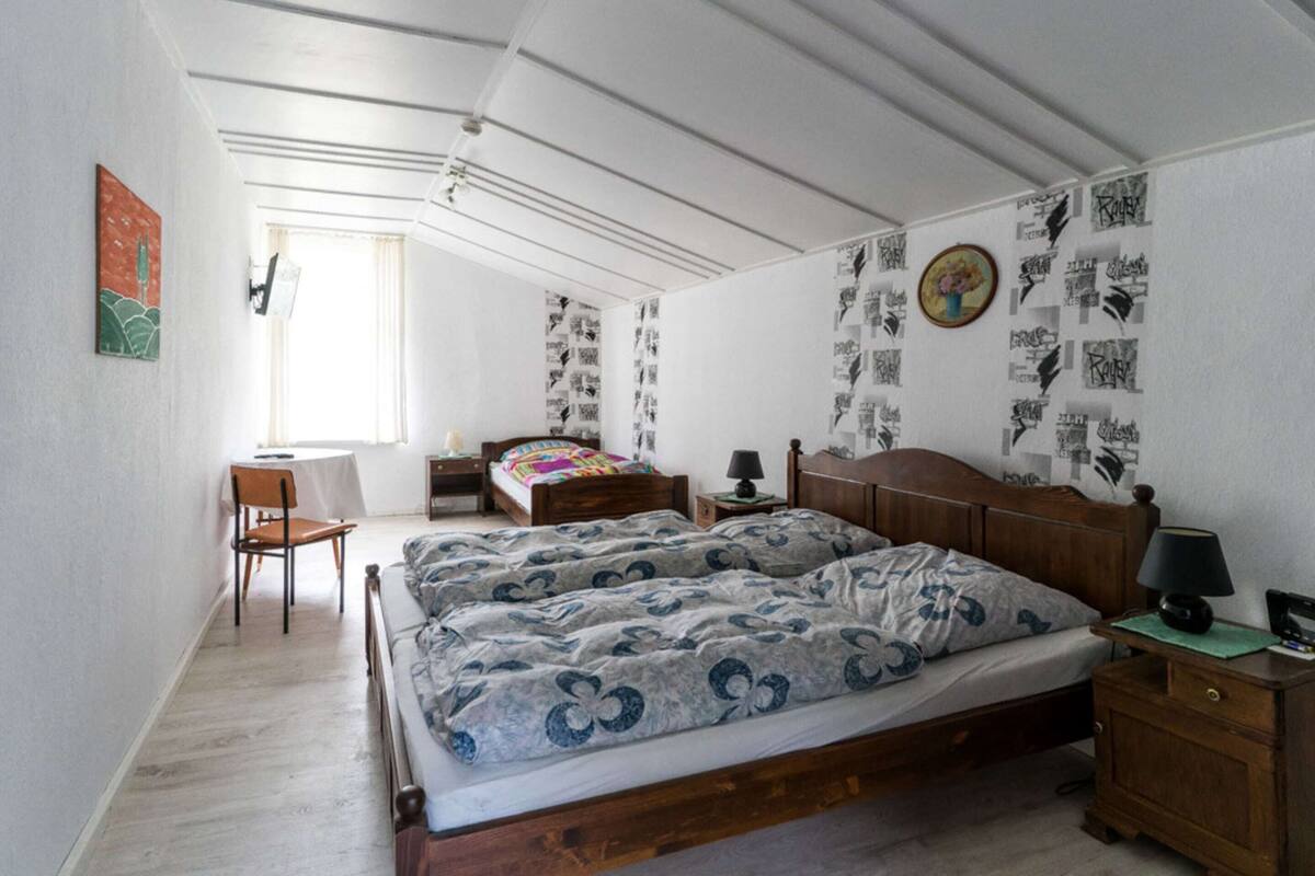 Ein Schlafzimmer mit einem großen Bett, einem Schreibtisch und einem Sessel. Mehrere Wanddekorationen und ein Fenster sorgen für natürliches Licht.