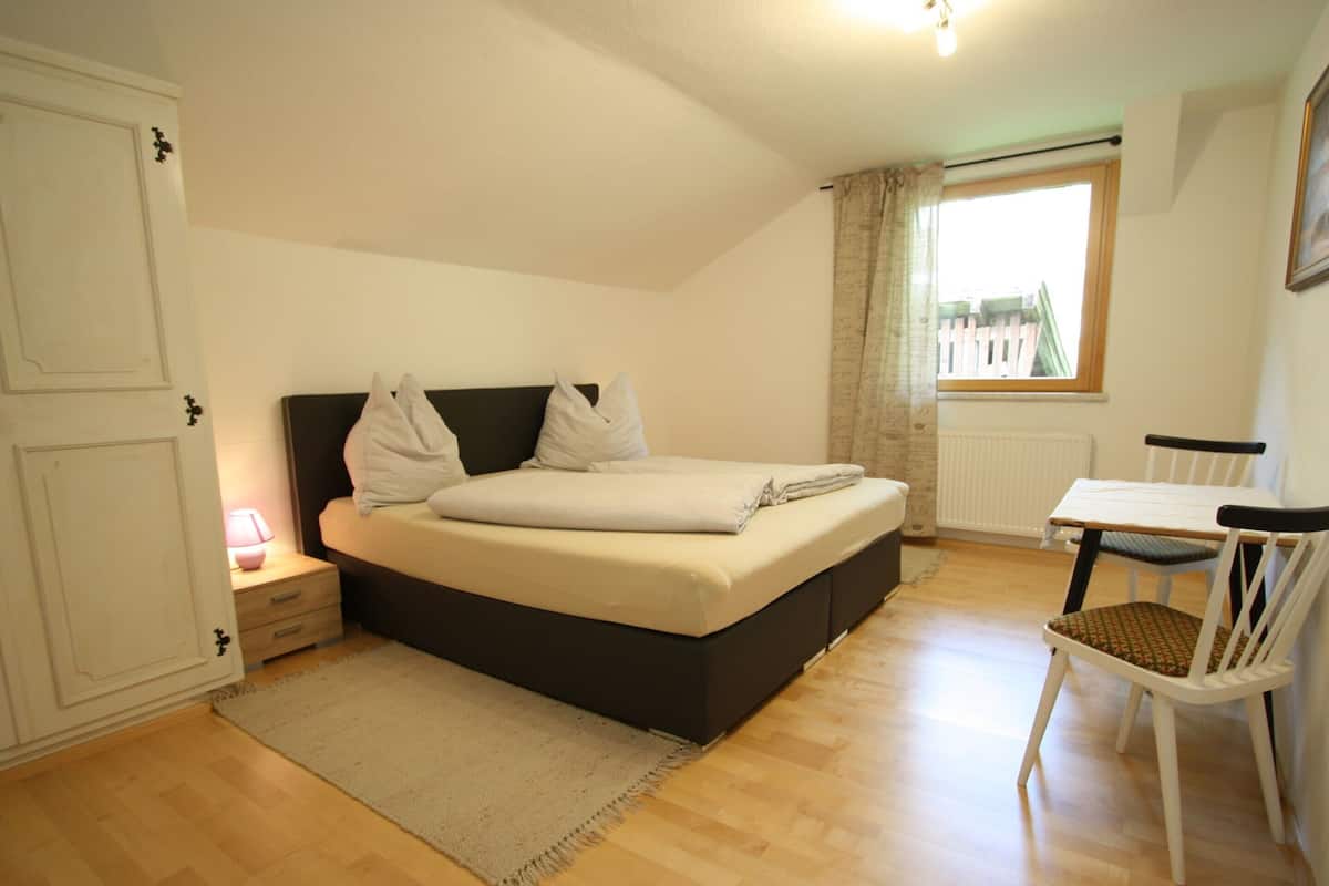 A bedroom with a bed, bedside table, chair, and a window with curtains.