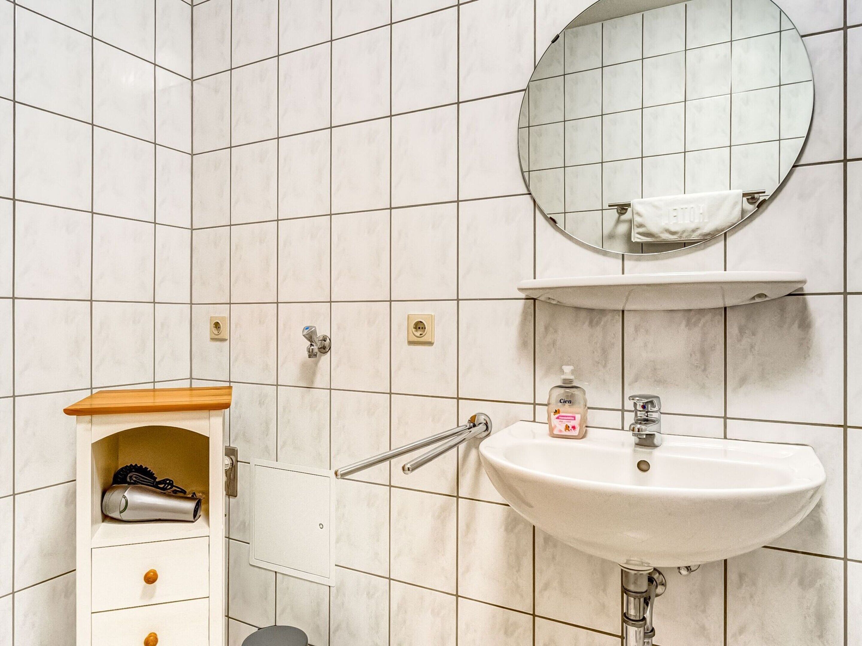 A bathroom with a white sink, a mirror, and a towel rack. There is a shelf with a hairdryer and a bottle of hand soap.