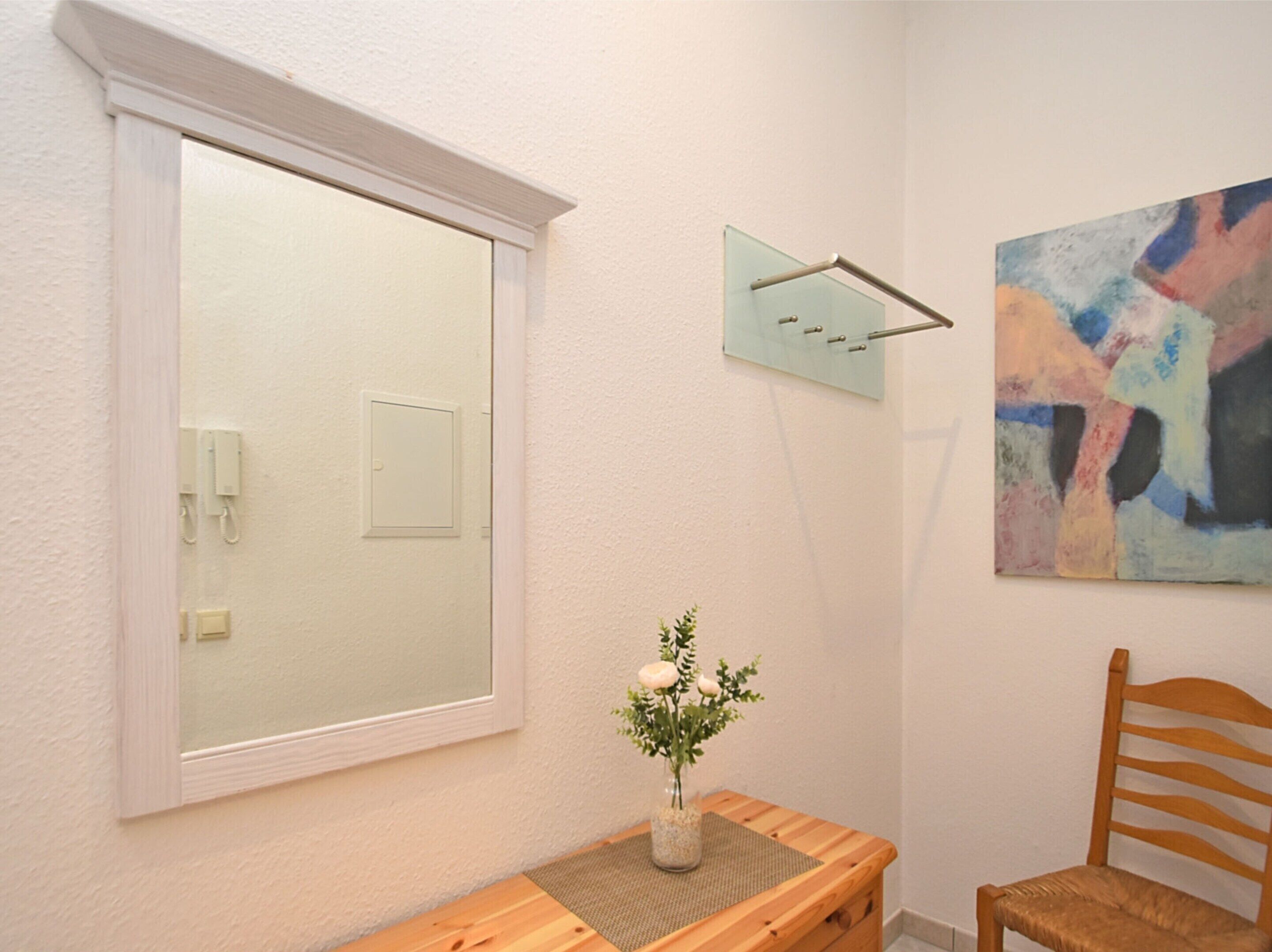 A room with a large window, a wooden chair, a small table with a vase of flowers, and a wall-mounted light fixture.
