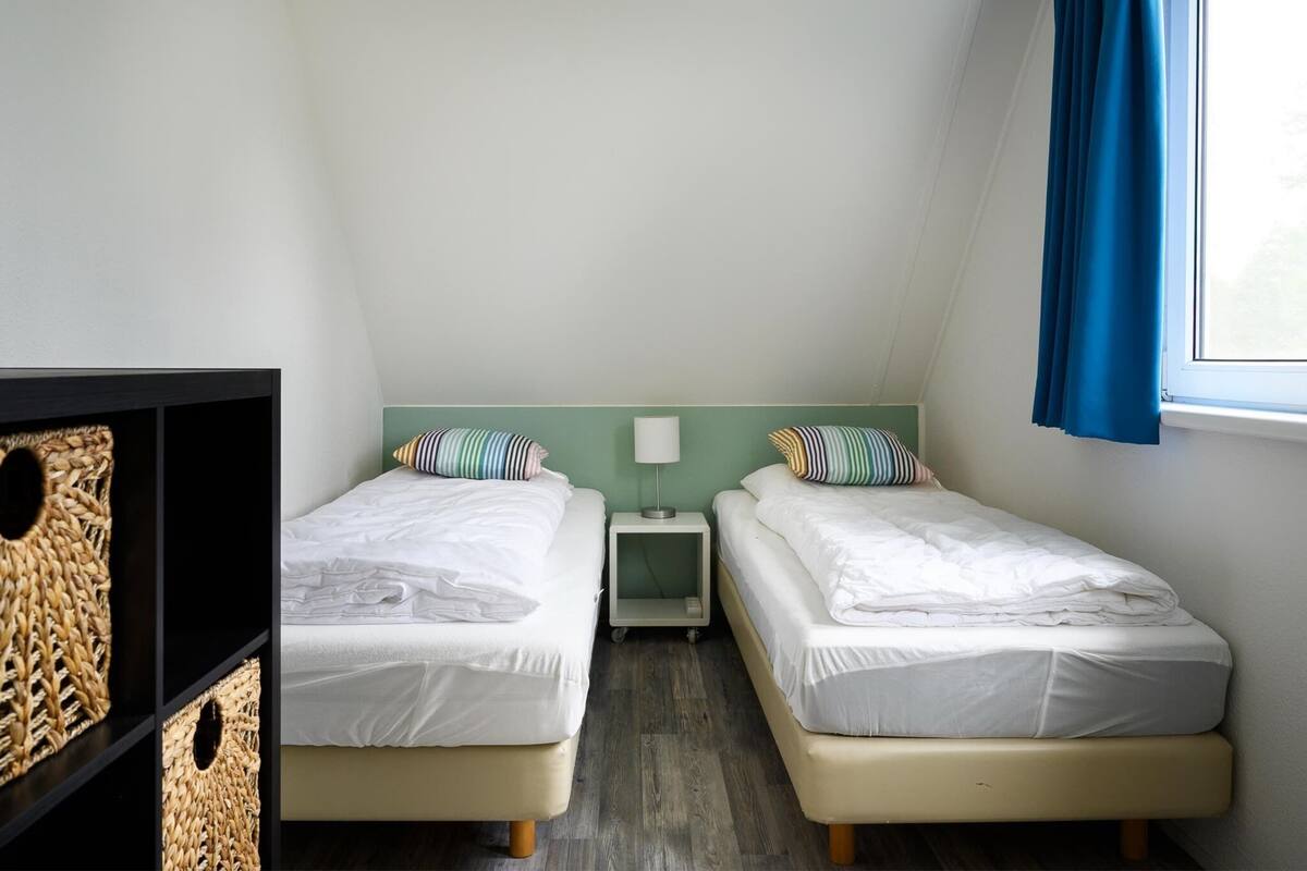 A small, well-lit room with two single beds, a nightstand, a lamp, and a window with blue curtains.