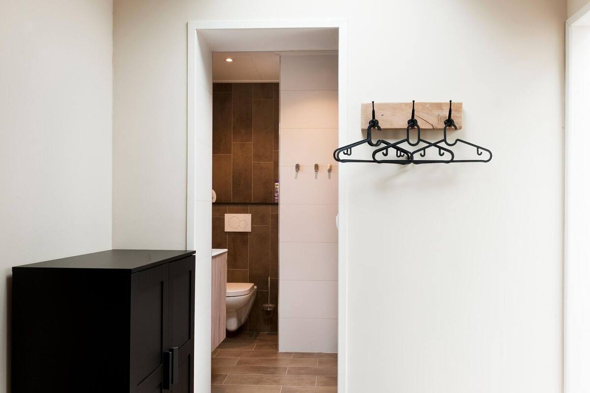 A narrow hallway with a black cabinet on the left, a white wall with a black metal coat rack on the right, and a view into a bathroom with a toilet and tiled walls.
