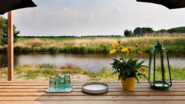 A wooden deck with a view of a river, surrounded by greenery and a clear sky. - House With Sauna, Outdoor spa and Pedal Boat in Vlagtwedde (Vlagtwedde)