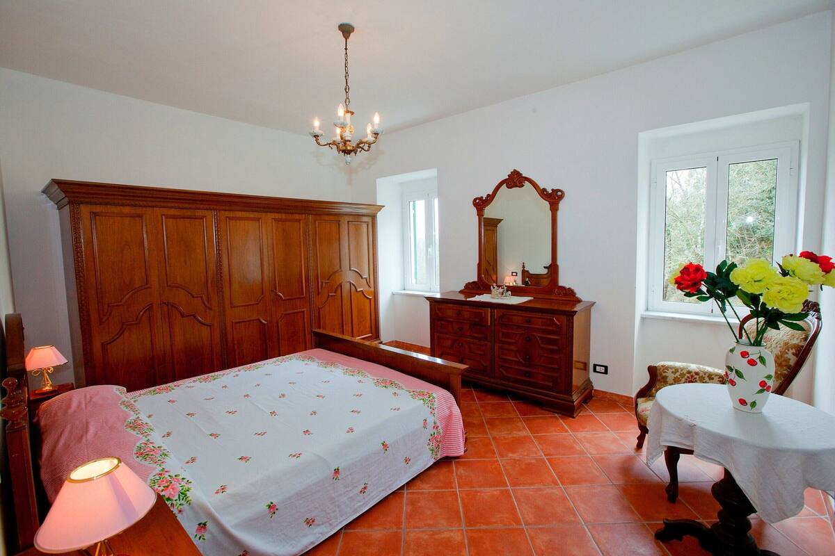 A bedroom with a wooden wardrobe, a bed with floral bedding, a dresser, a mirror, a chair, and a table with a vase of flowers.