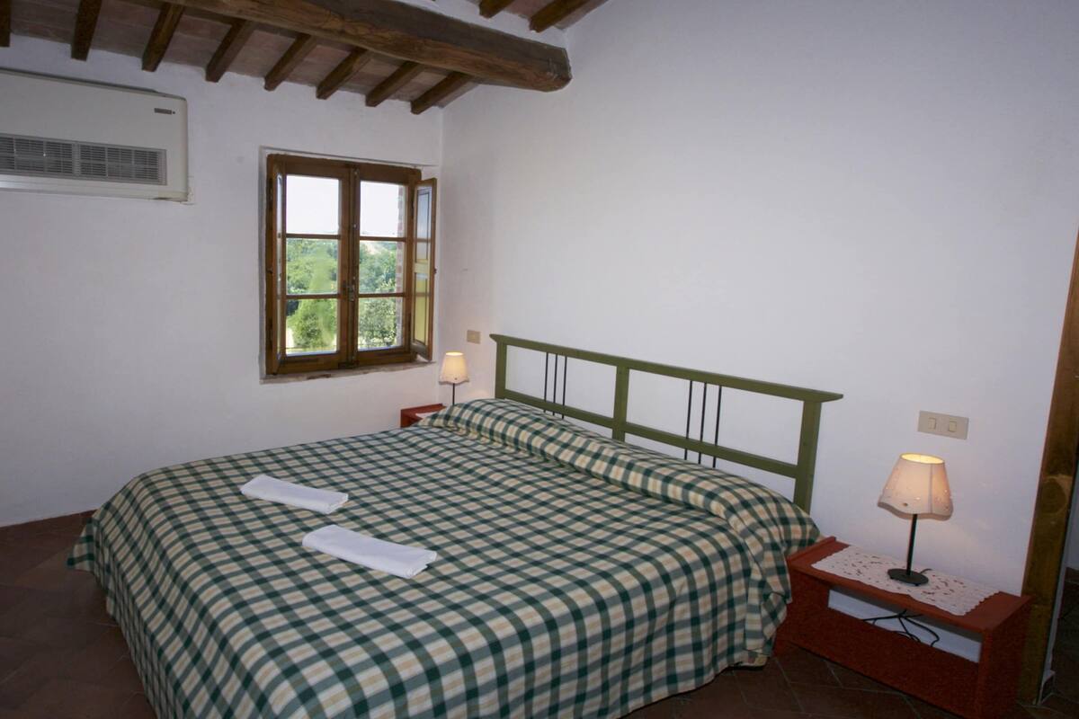 A bedroom with a bed, a window, a bedside table with a lamp, and a wall-mounted air conditioner.
