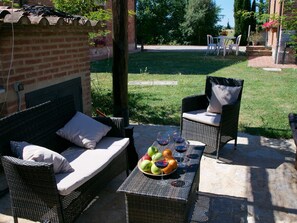 An outdoor seating area with wicker furniture, a table with fruit and wine glasses, and a brick wall in the background. - Lovely Farmhouse in Tuscany With Panoramic View (Castelnuovo Berardenga)