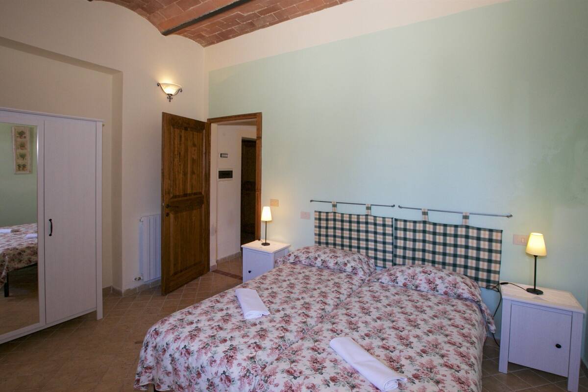A bedroom with a bed, two bedside tables with lamps, a wardrobe, and a door leading to another room.