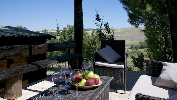 A patio with wicker furniture, a table with wine glasses and fruit, and a view of the countryside.