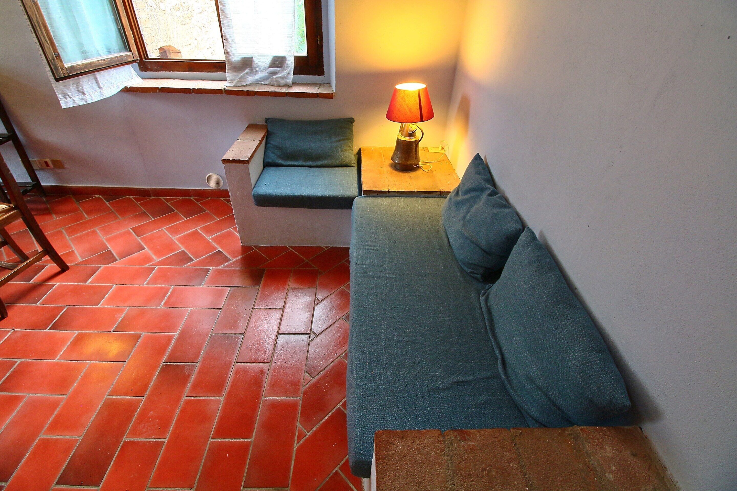 Une pièce avec un sol carrelé de tuiles rouges, un banc en bois, un canapé avec des coussins et une table avec une lampe.