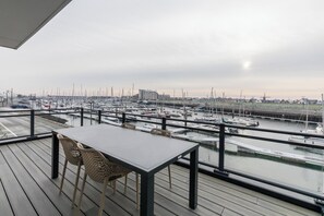 Ein Balkon mit Tisch und Stühlen mit Blick auf einen Yachthafen, in dem zahlreiche Boote und ein entferntes Gebäude zu sehen sind.
