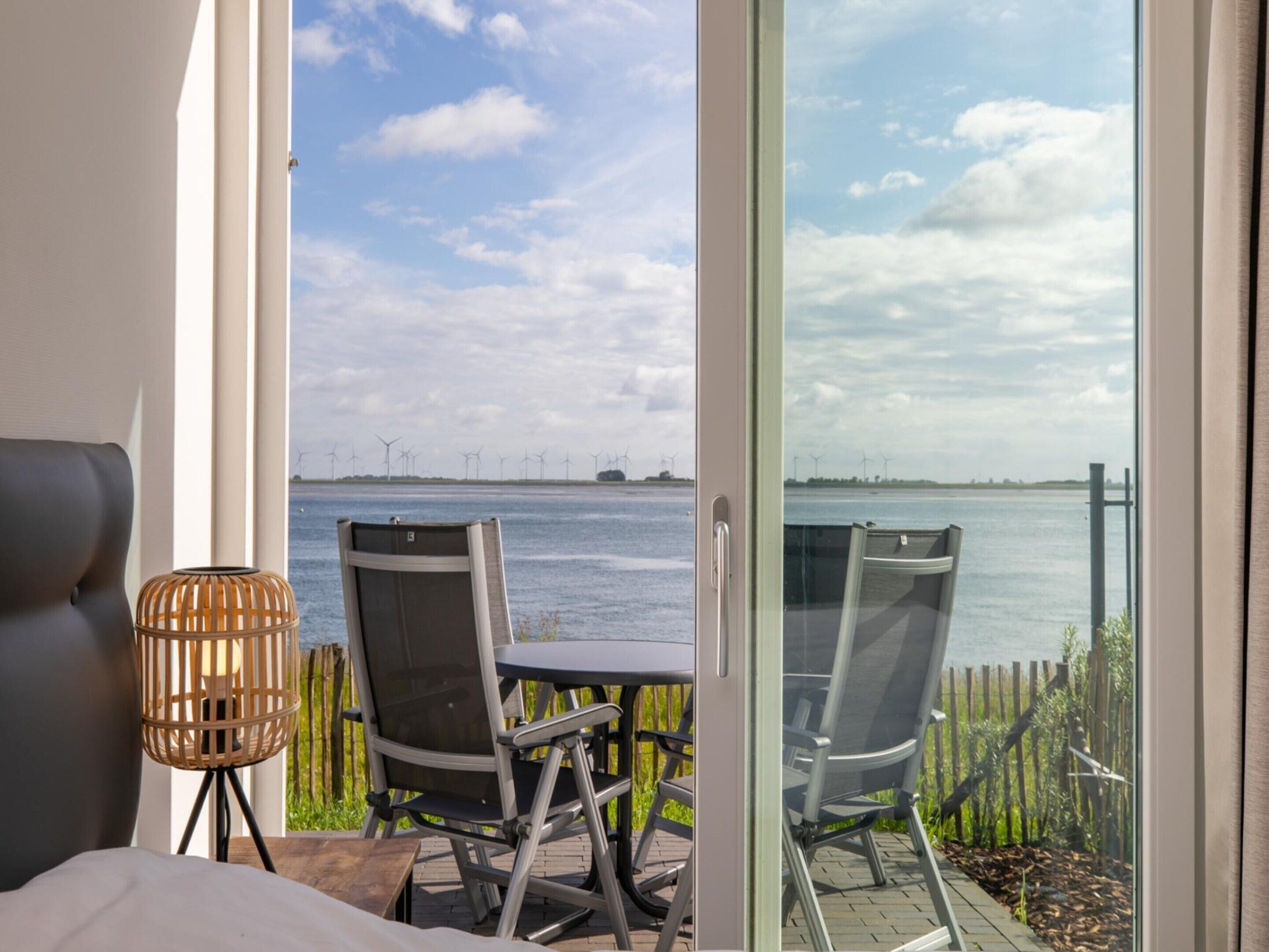 A room with a bed, a table, and two chairs facing a large window overlooking a body of water and wind turbines.