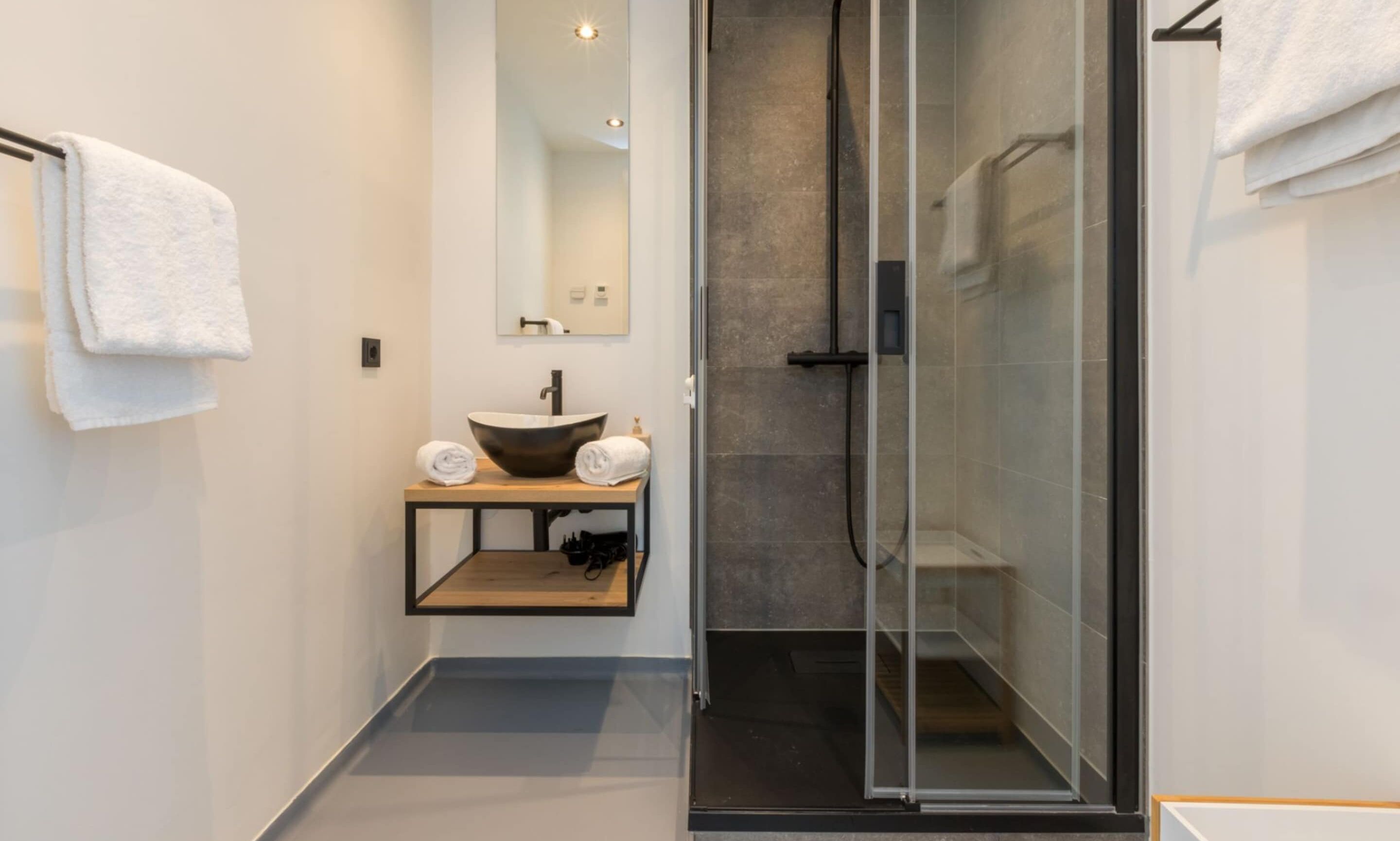Un baño moderno con ducha envuelta en vidrio, tocador de madera con un lavamanos grande y un espejo sobre el lavamanos.