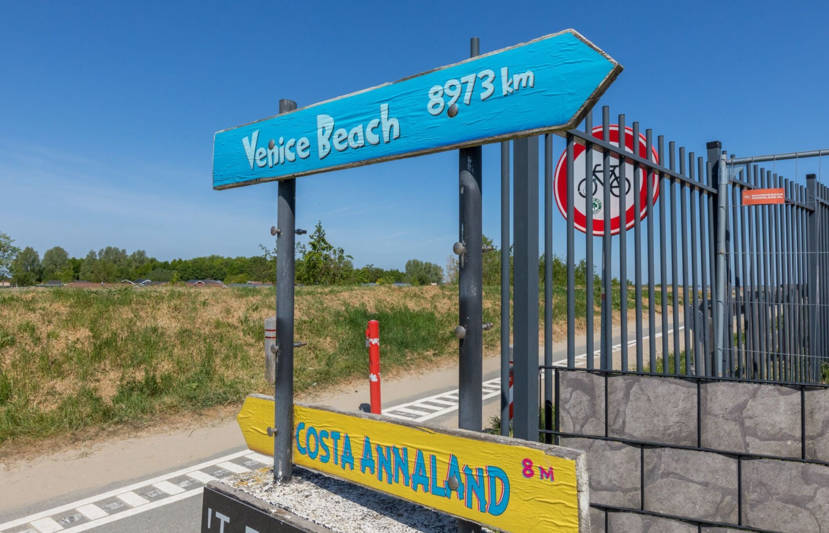Un cartel con 'Venice Beach' y '8973 km' indicando la distancia, un símbolo de bicicleta y un cartel de 'Costa-Annaland' con un indicador de distancia.