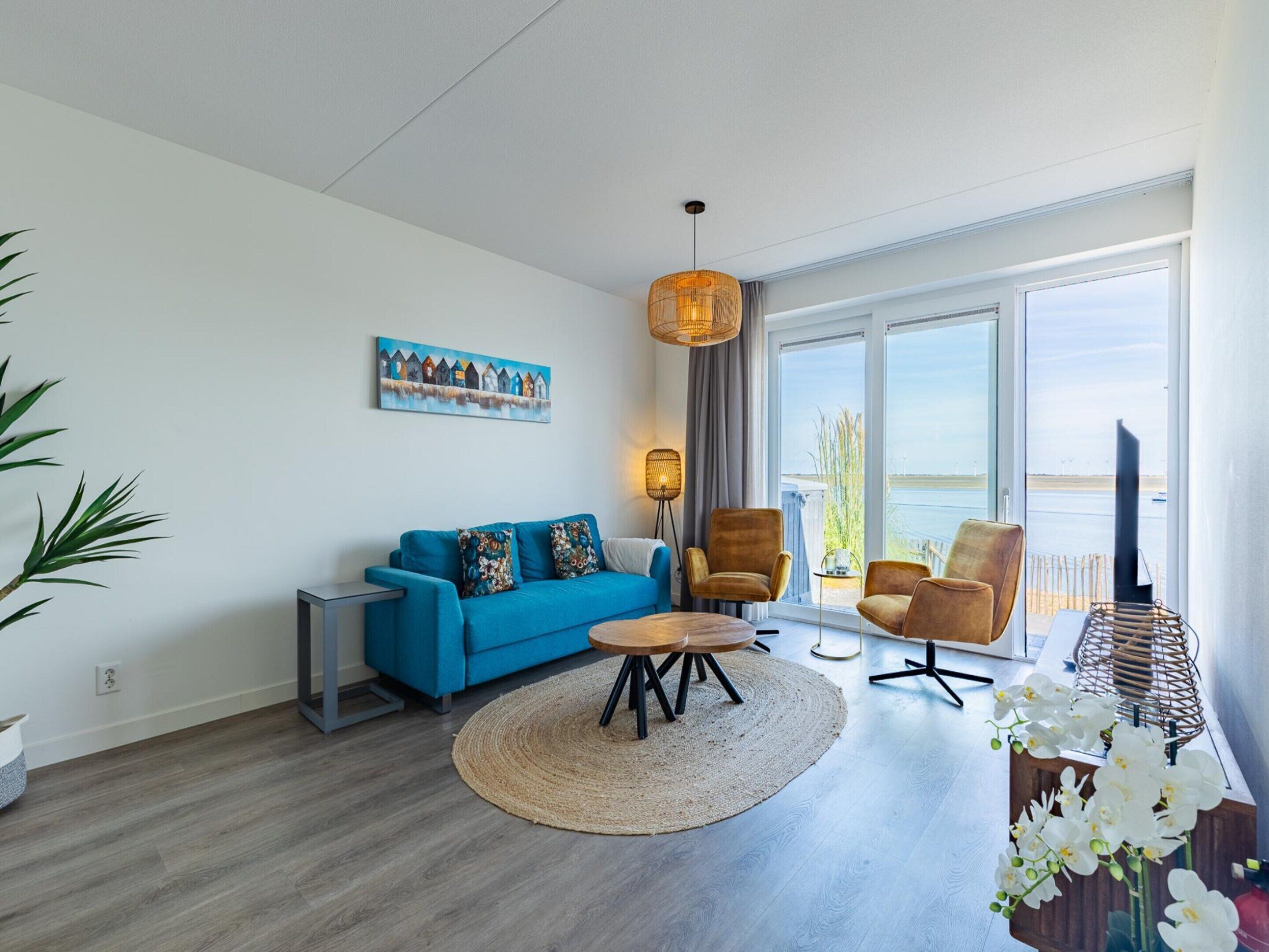 A modern living room with a blue sofa, two armchairs, a coffee table, and a large window offering a view of the water.