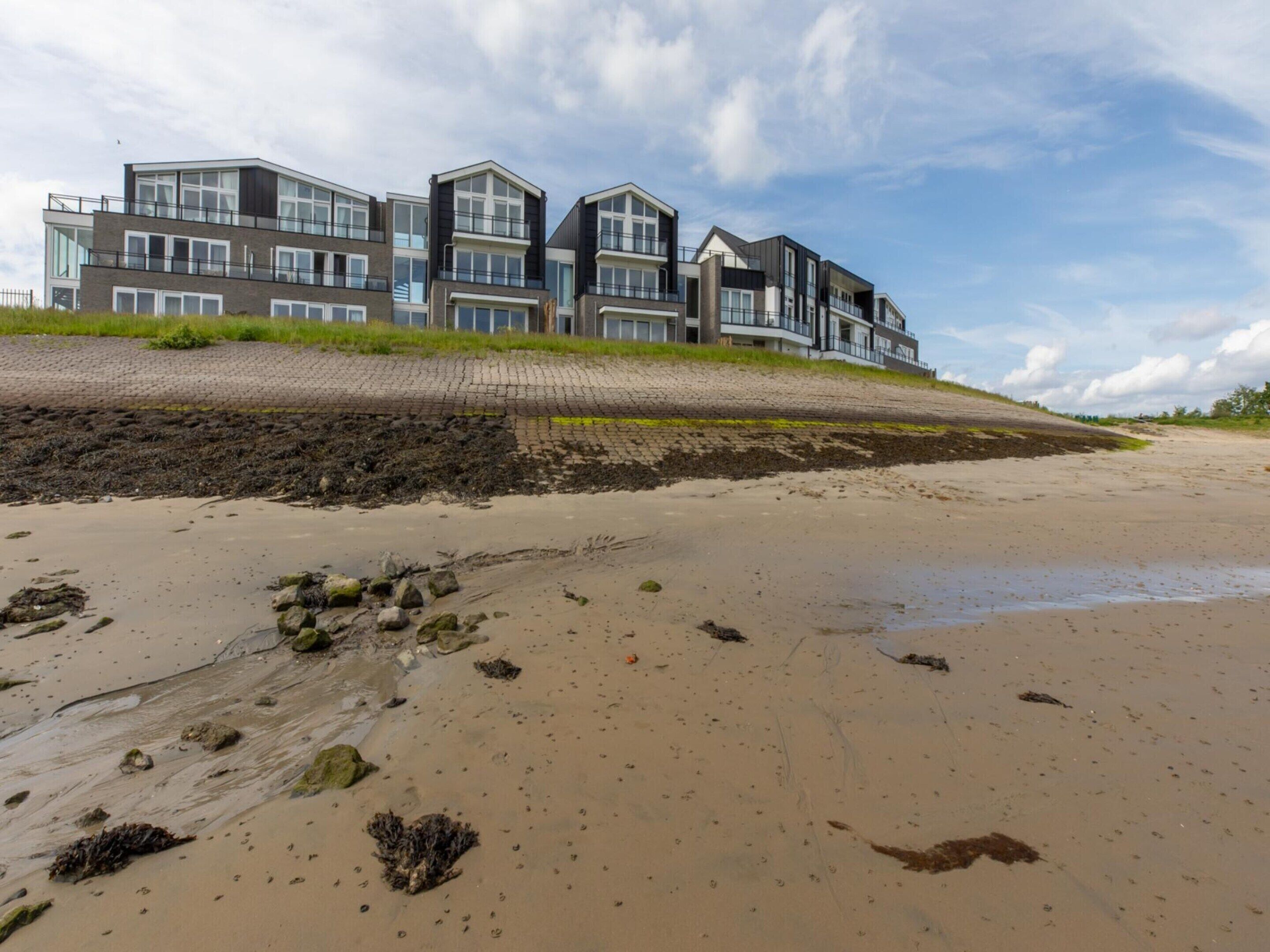 Un immeuble moderne à plusieurs étages, doté de grandes fenêtres et de balcons, situé sur une plage de sable fin, le long d’une côte rocheuse.