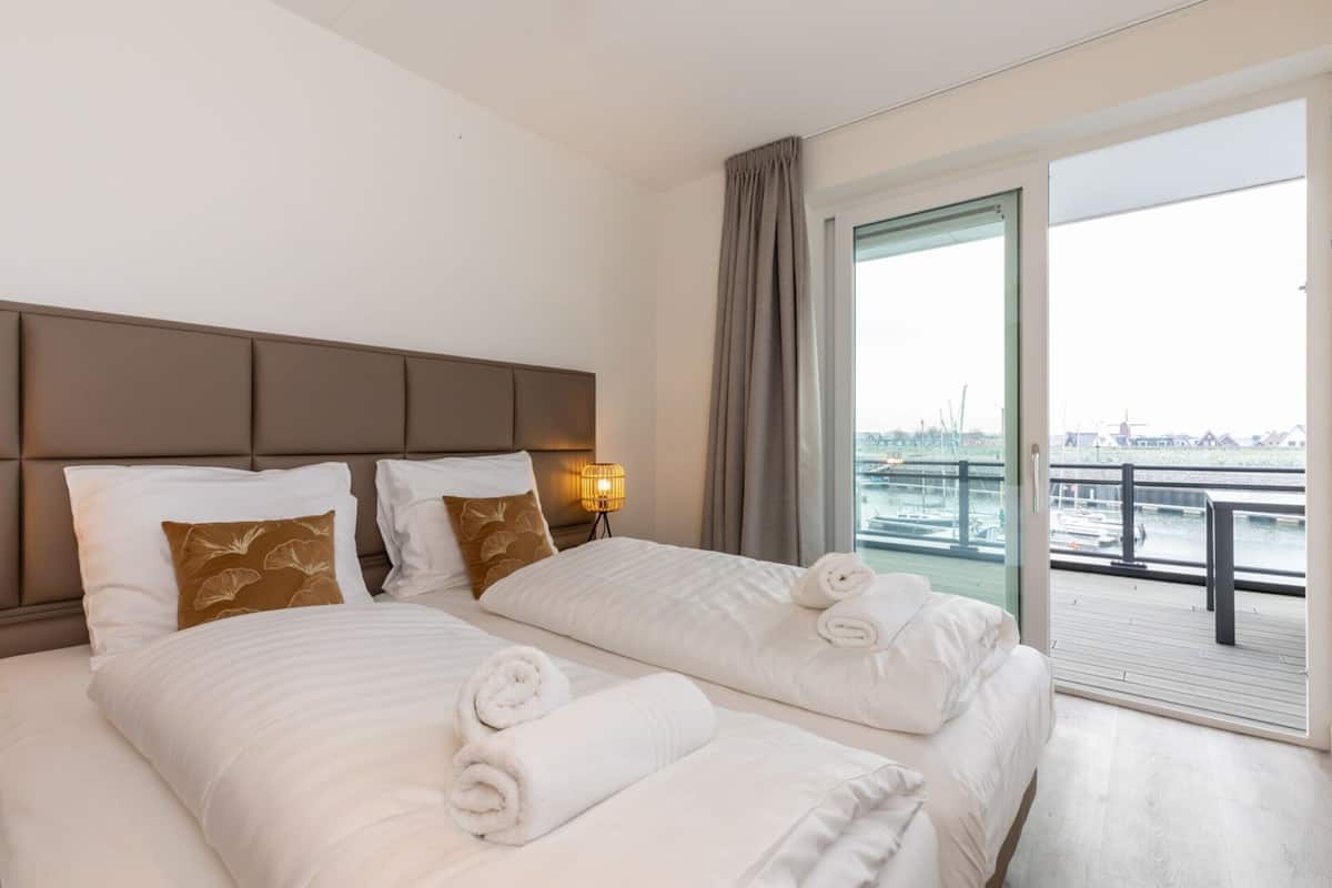 Une chambre d’hôtel avec un grand lit, deux oreillers, une table de chevet avec une lampe et une vue sur un plan d’eau, accessible par une porte coulissante.