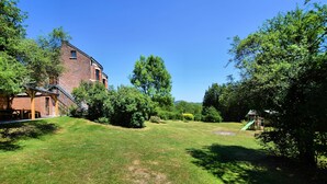 A grassy backyard with a brick building, trees, and a wooden play structure. - Cozy Cottage in Durbuy With Garden (Durbuy)