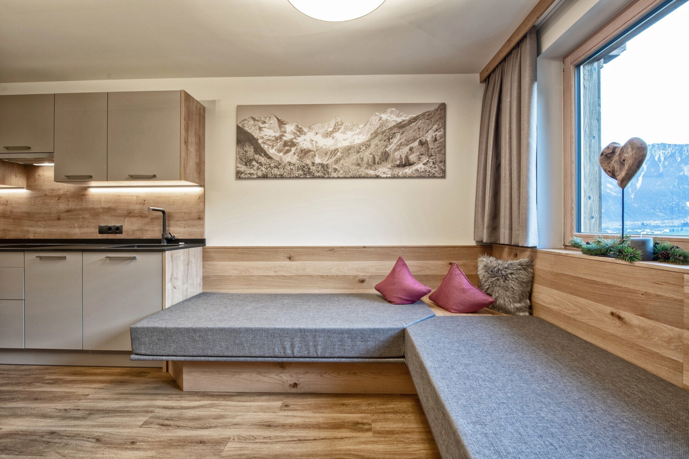 A modern kitchen with wooden cabinets, a built-in bench with cushions, a large window with a mountain view, and a framed landscape painting on the wall.