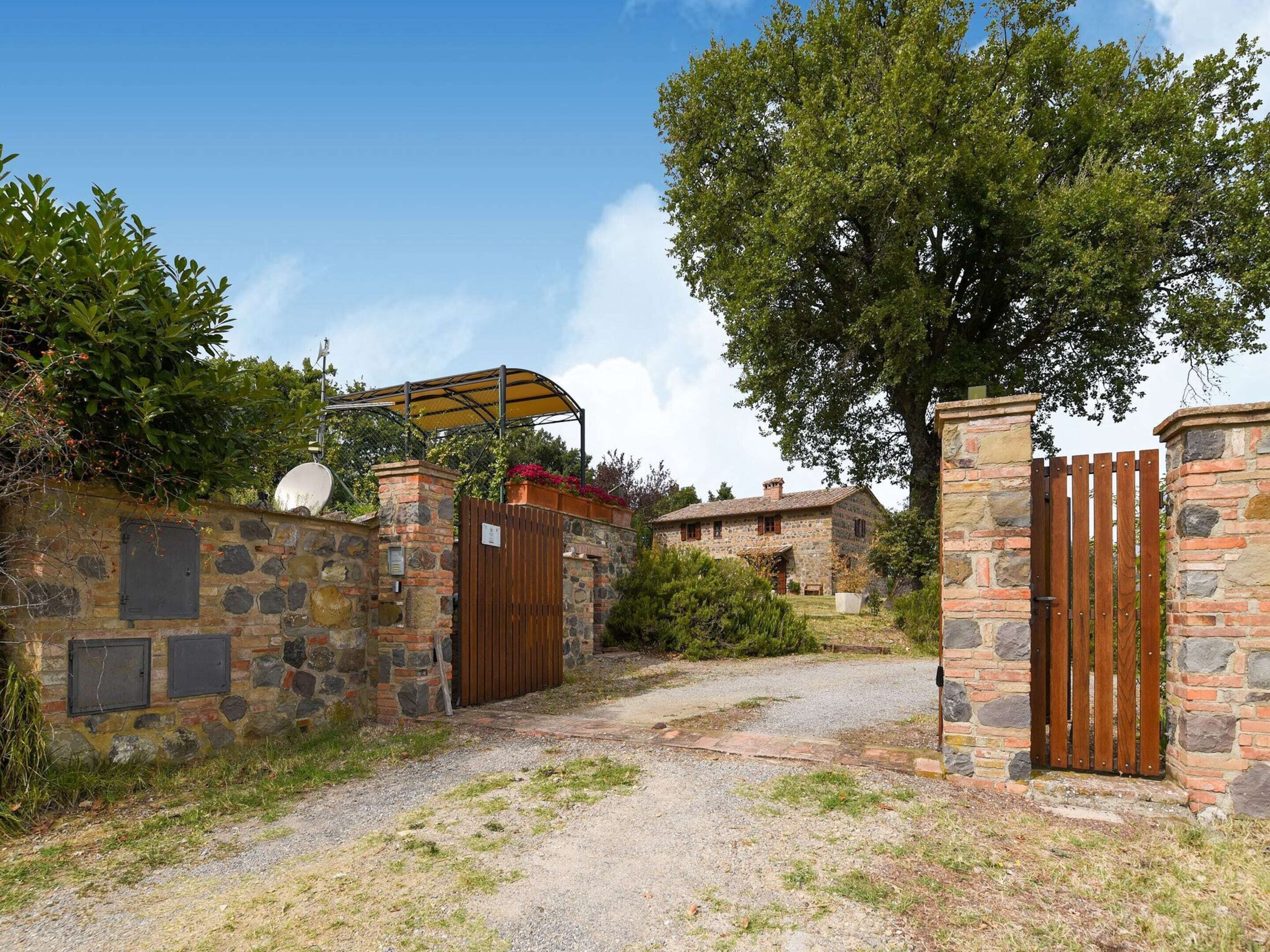 A rustic stone building with a wooden gate, a covered area with a satellite dish, and a gravel path leading to the entrance.