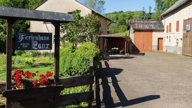 A sign for 'Ferienhaus Lauz' with a wooden bench and red flowers in the foreground, and a building with a sloped roof in the background. - Spacious Holiday Home in Ulmen Near the Centre (Ulmen)