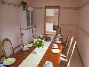 A dining room with a wooden table set for six, white chairs, a white cabinet with glass doors, and a view into the kitchen through an open doorway. - Spacious Holiday Home in Ulmen Near the Centre (Ulmen)