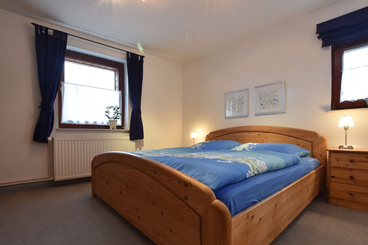 Een slaapkamer met een houten bed, een nachtkastje, een lamp, een raam met gordijnen en twee aan de muur hangende foto's.