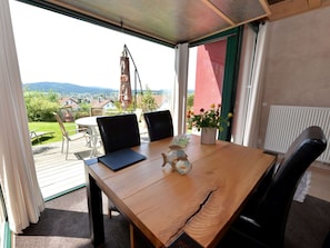 A wooden table with chairs, a laptop, a vase with flowers, and a view of a town and mountains through a window. - Tranquil Holiday Home in Blossersberg With Terrace (Viechtach)