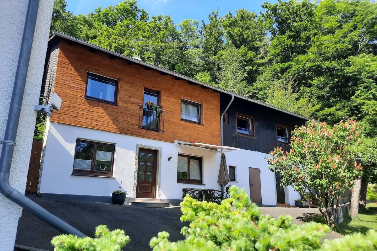 Ein zweistöckiges Haus mit weiß-brauner Fassade, einer Haustür und einer kleinen Terrasse mit Gartenmöbeln.