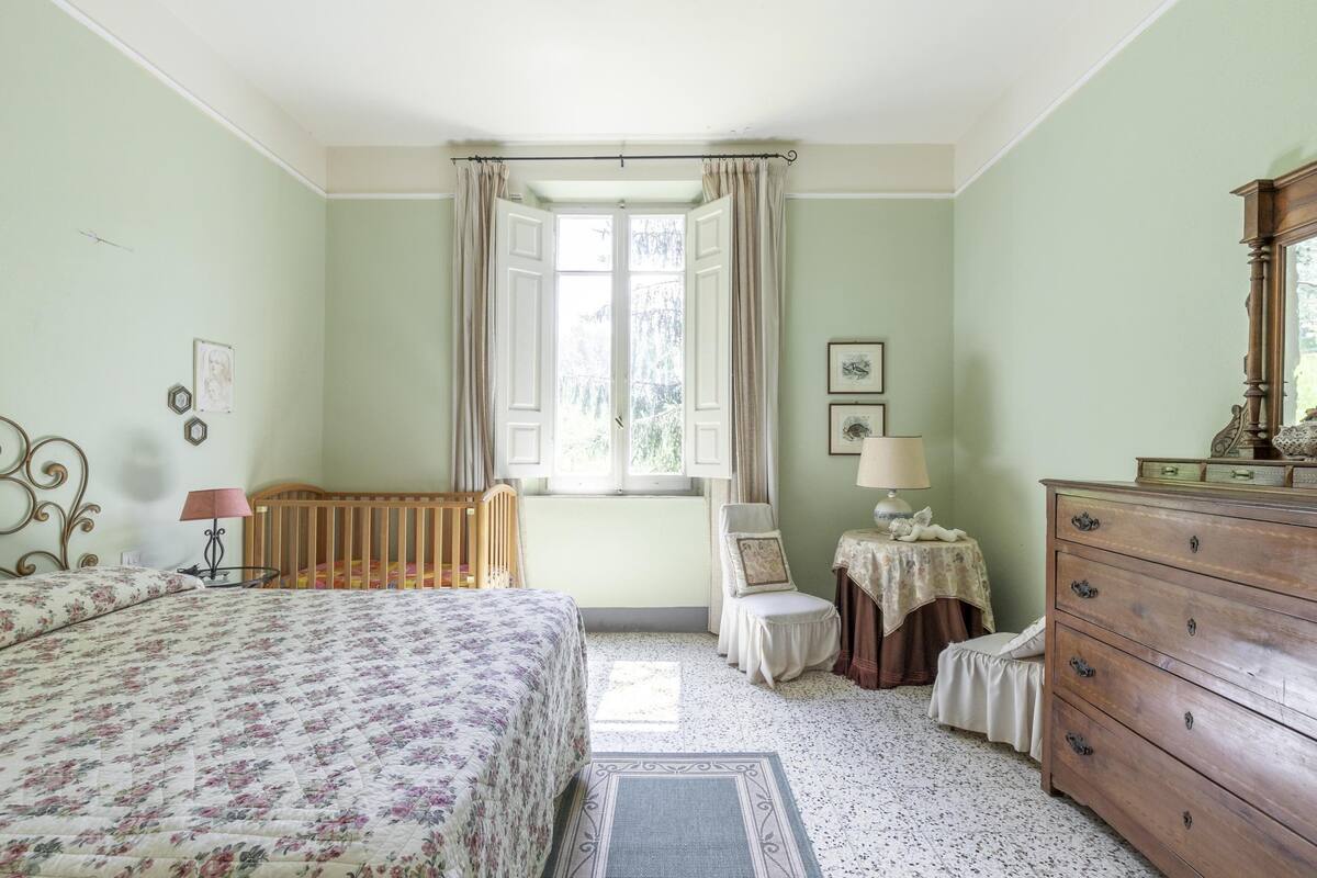 A bedroom with a bed, a wooden dresser, a chair, a small table, and a window with curtains.