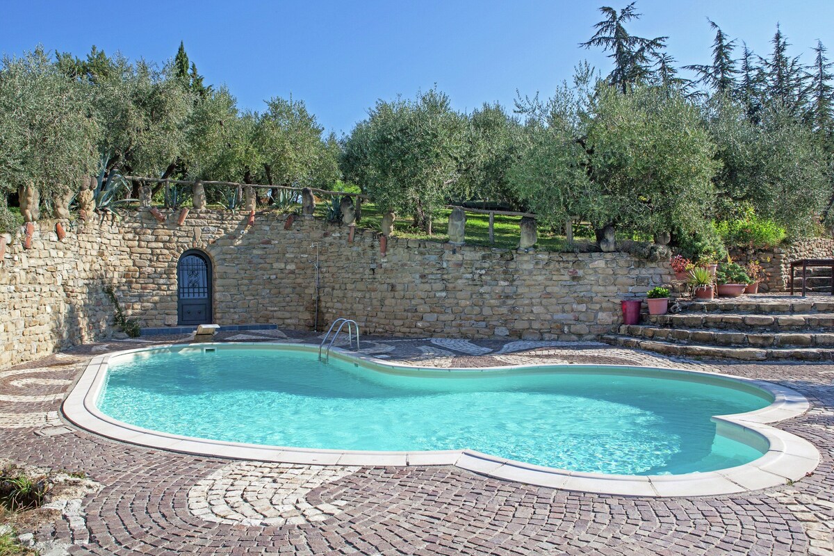 Una piscina con un muro de piedra y escalones que la bordean, rodeada de árboles y vegetación.