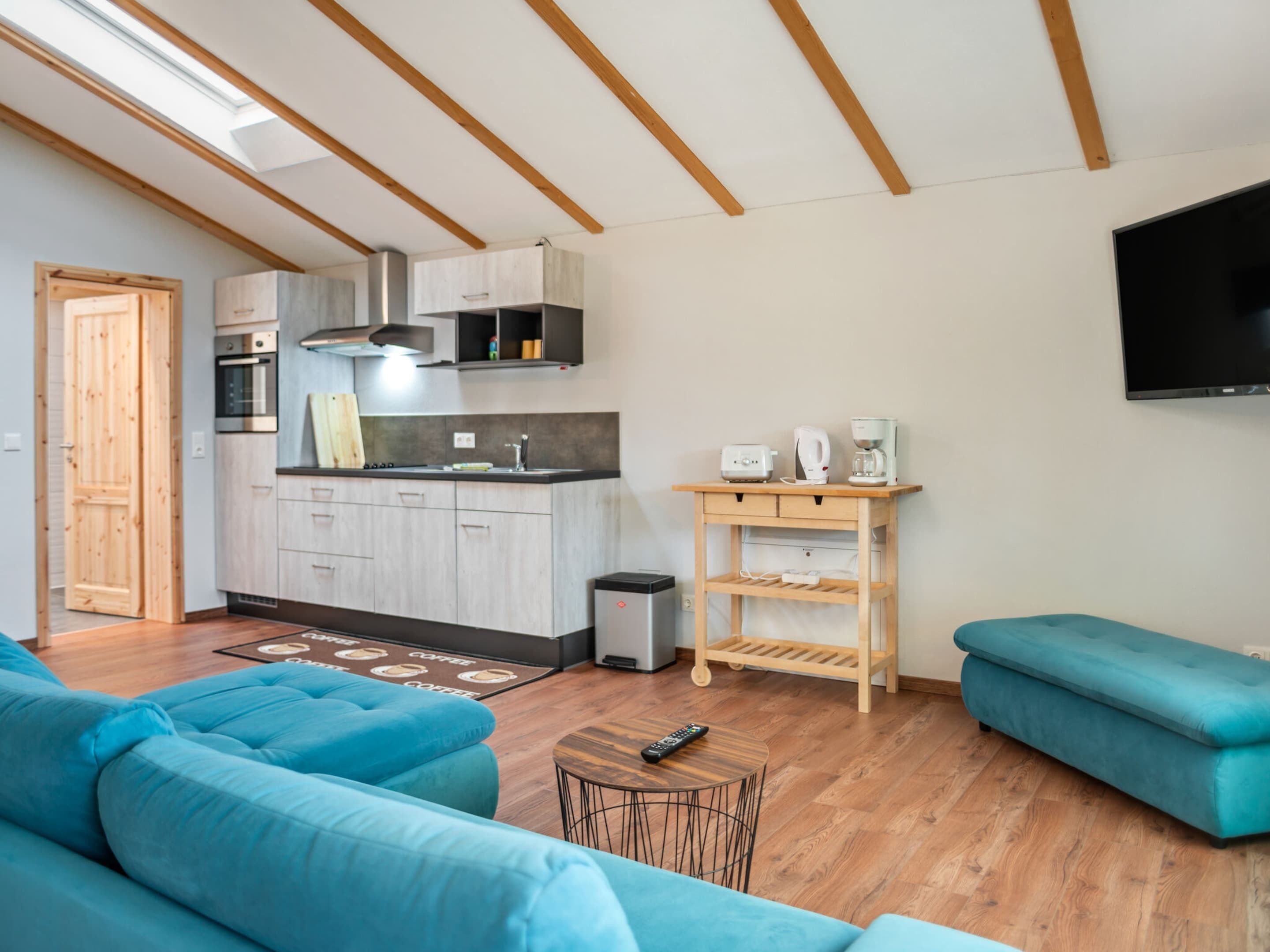 A modern living room with a sofa, a coffee table, and a TV. There is a kitchen area with cabinets and appliances.