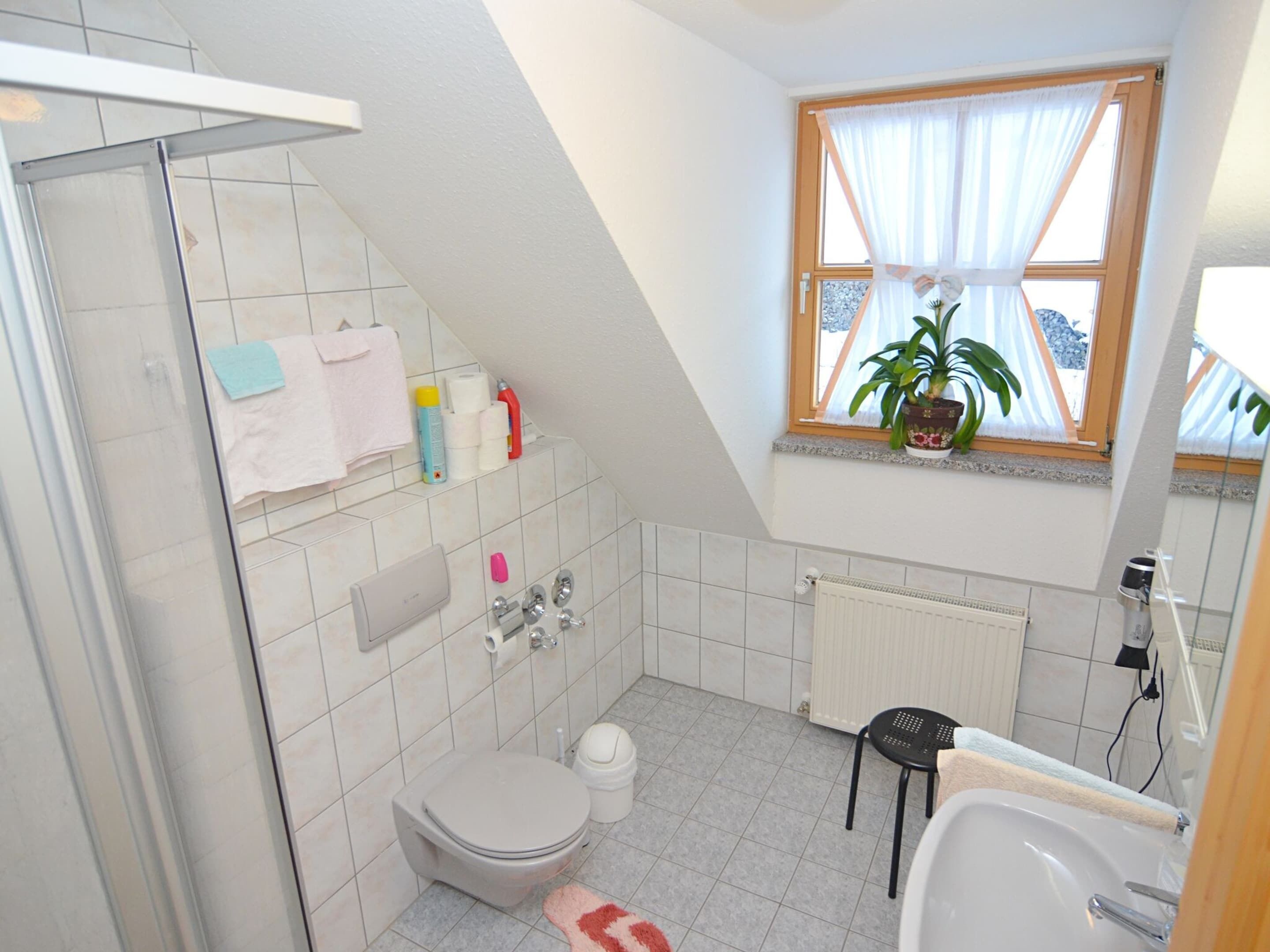 A small, modern bathroom with a shower, toilet, and sink. There is a window with a plant on the windowsill, and toiletries are visible on the wall.