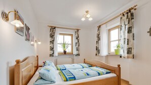 A bedroom with a wooden bed, two bedside tables, a nightstand, a window with patterned curtains, a radiator, and wall-mounted lighting. - Spacious Apartment With Sauna in Schonsee (Schoensee)
