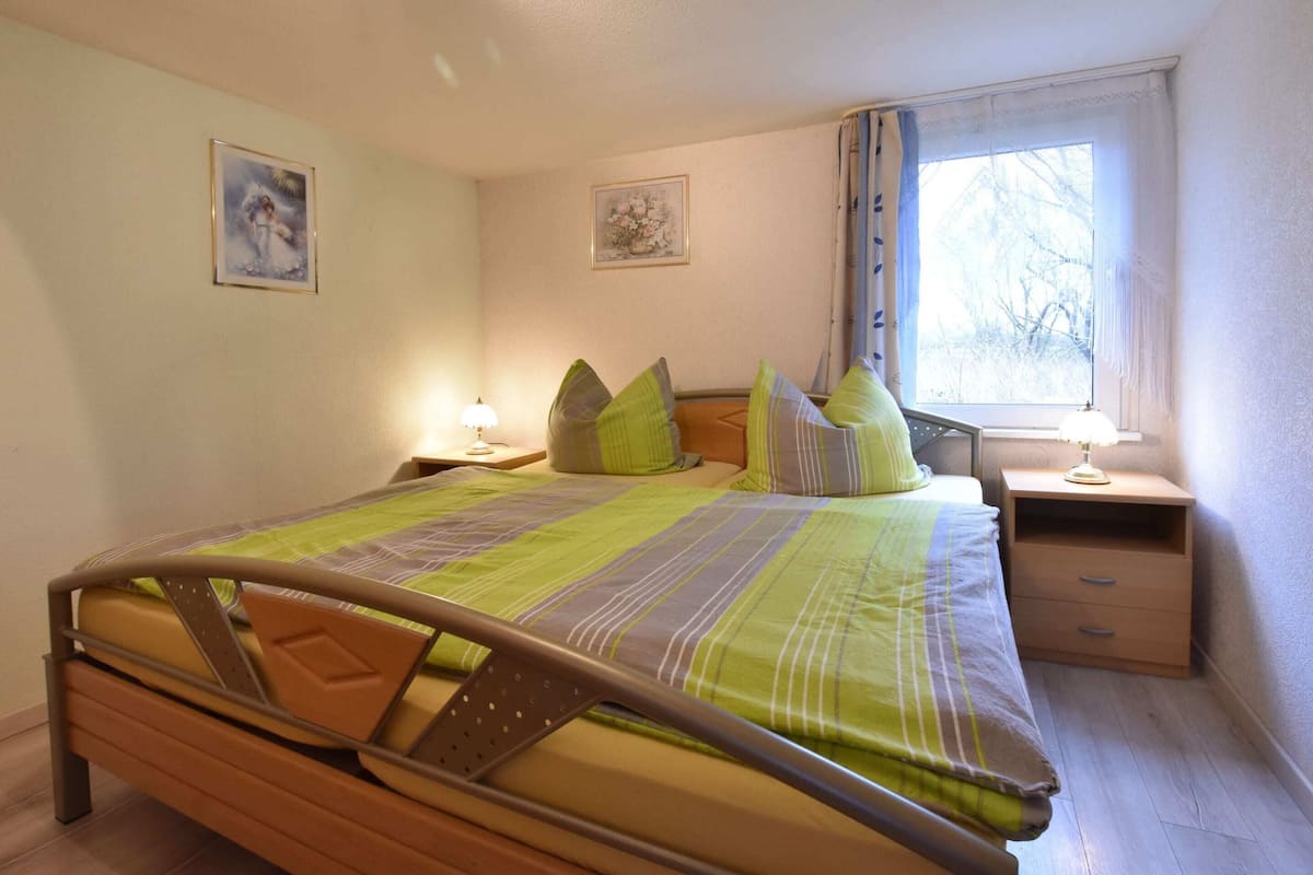 Une chambre à coucher comprenant un lit, deux tables de chevet, une fenêtre avec des rideaux et deux tableaux accrochés au mur.