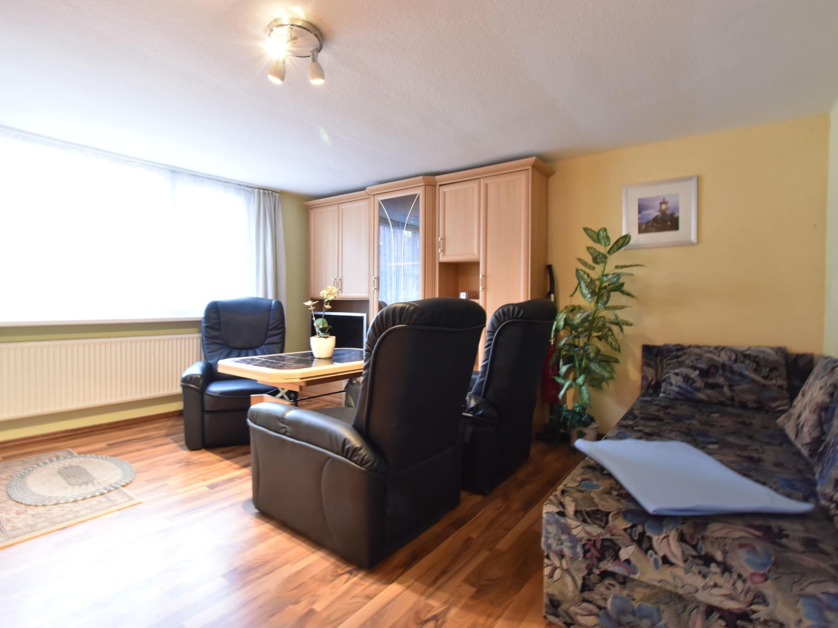 A living room with a sofa, two armchairs, a coffee table, and a potted plant. There is a window with curtains, a radiator, and a ceiling light fixture.