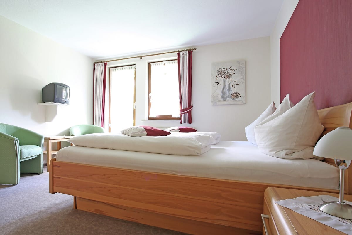 Une chambre à coucher avec un lit en bois, une chaise verte, un téléviseur posé sur une étagère et une fenêtre avec des rideaux.