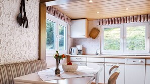 Une cuisine chaleureuse avec un coin repas, dotée d’un plafond en bois, de meubles blancs et d’une fenêtre donnant sur un paysage verdoyant.