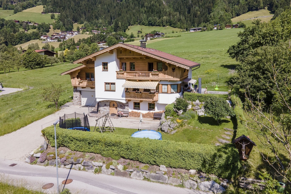 A two-story chalet with a balcony, surrounded by a lush green landscape and a clear sky.