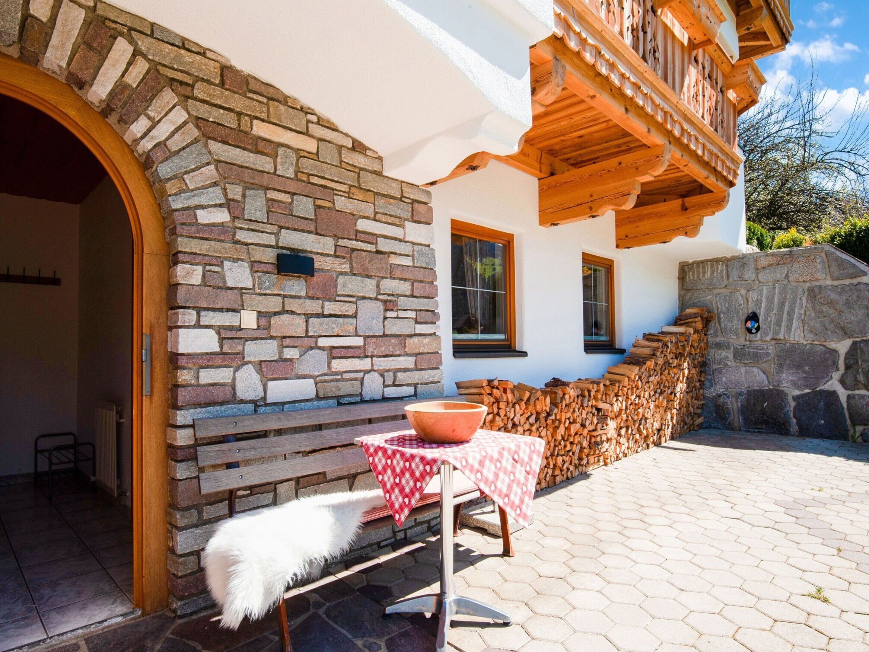 A stone exterior wall, a wooden bench, a table with a checkered tablecloth, and stacked firewood.