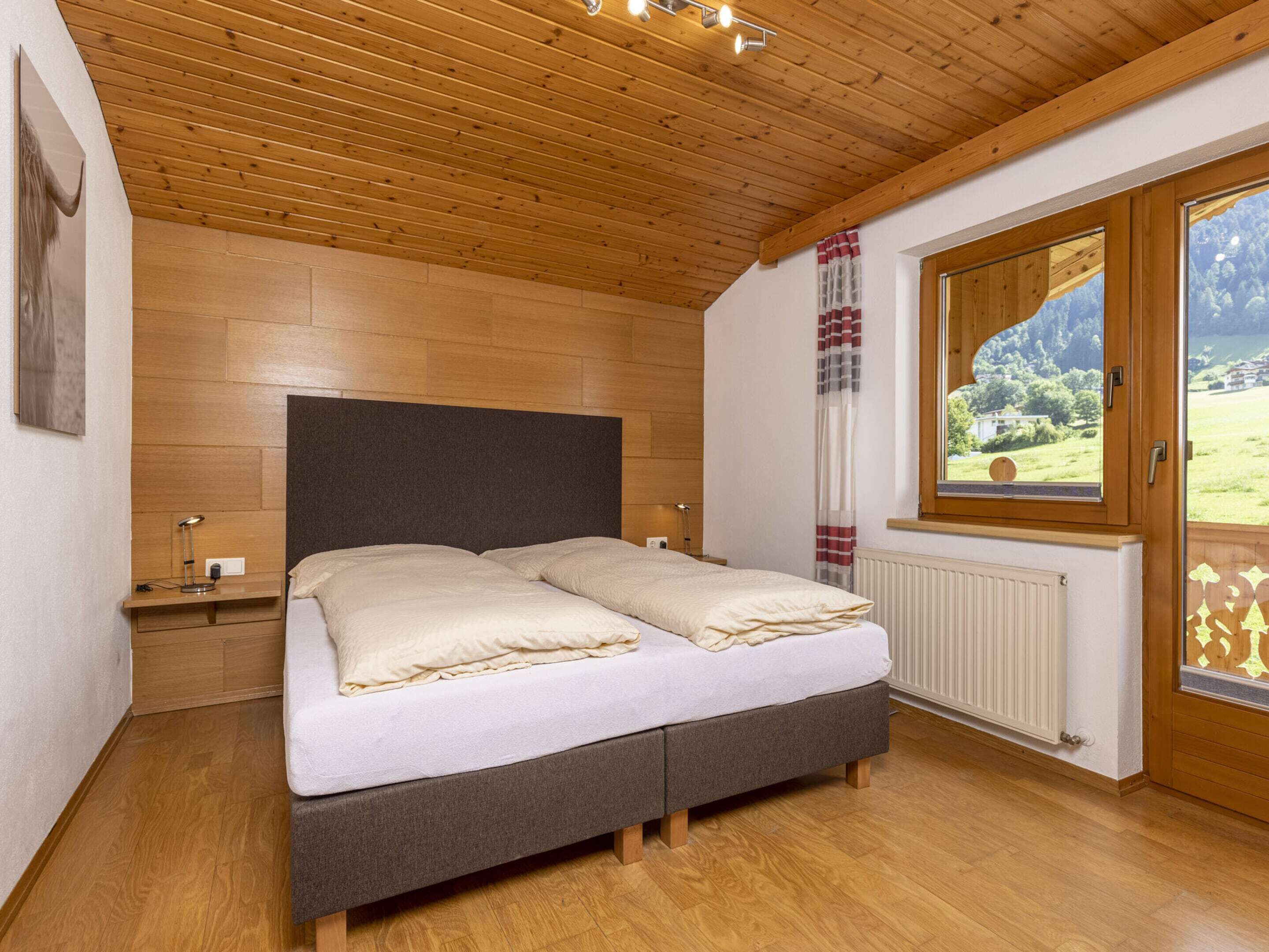 A bedroom with a wooden ceiling, a bed with a grey headboard, a nightstand, a window with a view of the outdoors, and a radiator.