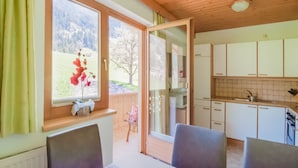 A dining area with a table and chairs, a window with a view of a mountain and flowers, and a kitchen with cabinets and a sink. - Apartment With Panoramic View (Ramsau im Zillertal)