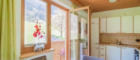 A dining area with a table and chairs, a window with a view of a mountain and flowers, and a kitchen with cabinets and a sink.